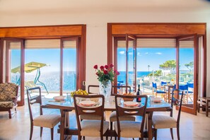 A dining area with a table set for four, a vase with flowers, and a view of the sea through large windows.