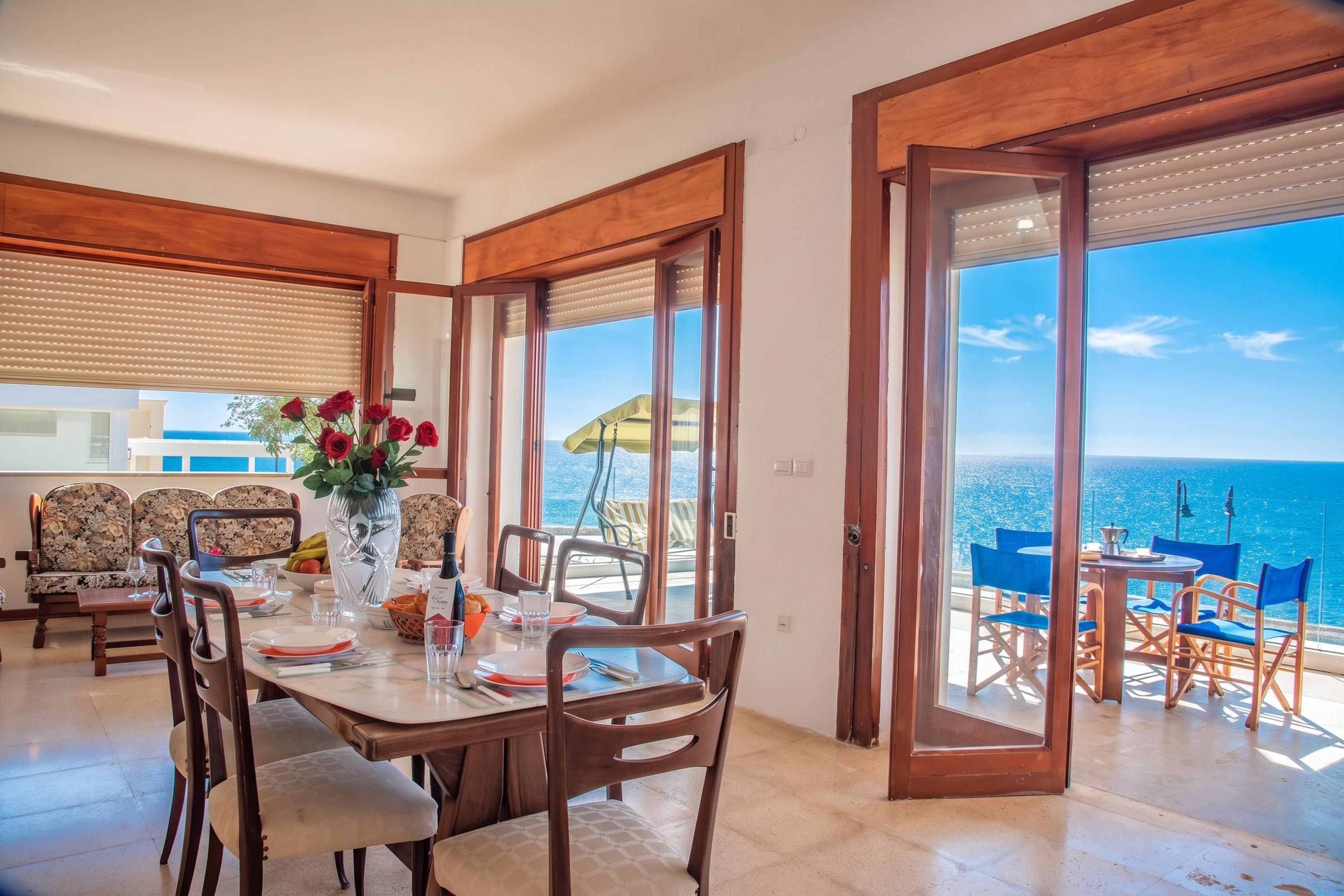 A dining area with a table set for four, a vase with red flowers, and a view of the sea through large glass doors.