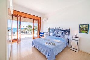 A bedroom with a bed, a nightstand, a window with a view of the sea, and a painting on the wall.
