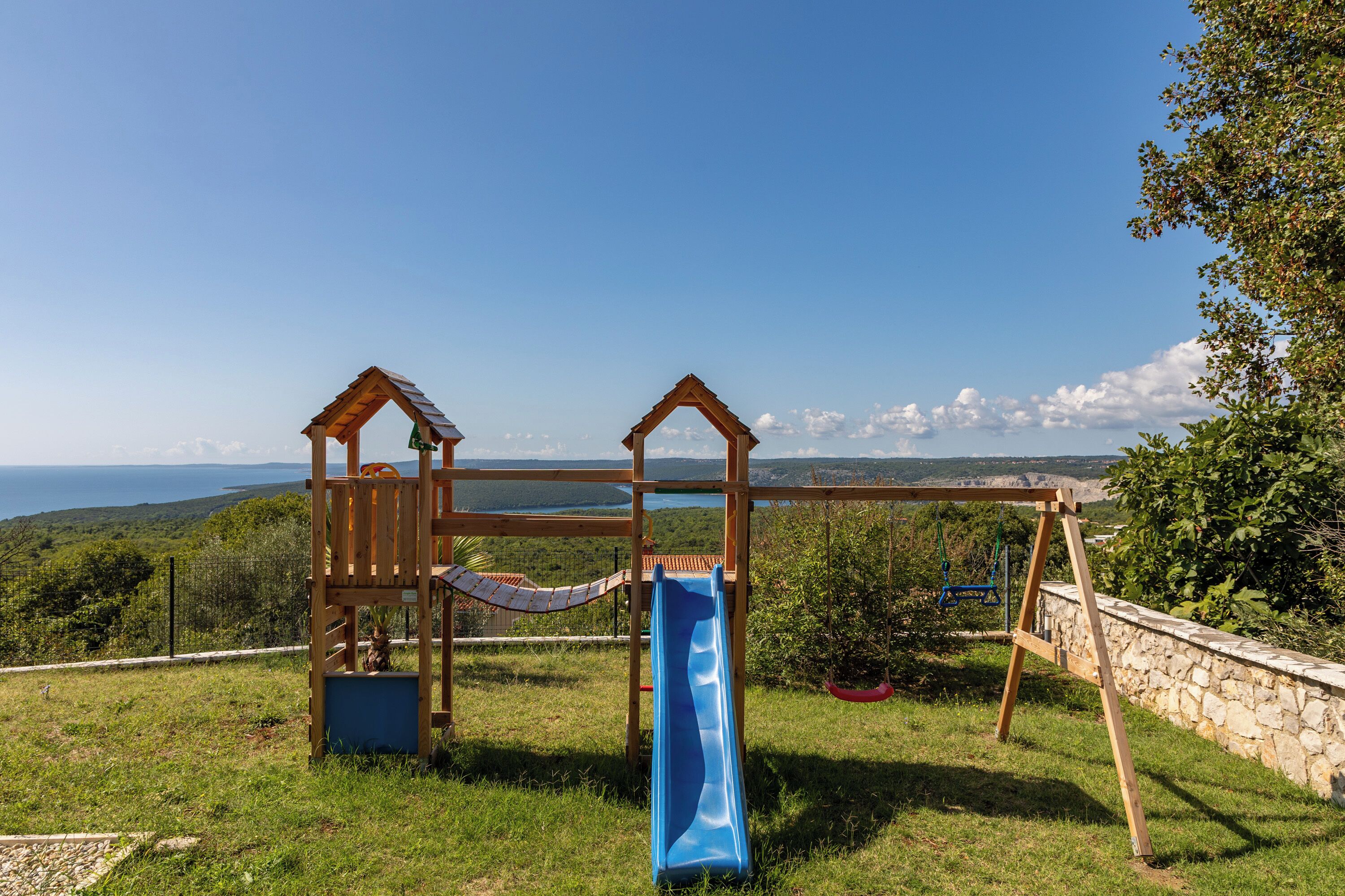 A playground with a slide and swings, set against a scenic backdrop of hills and a body of water.
