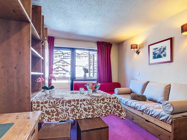 Una habitación acogedora con un sofá de madera, una mesa con mantel estampado y una ventana con cortinas.