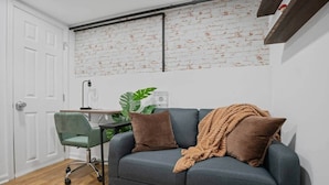 Una habitación con un sofá gris, dos cojines marrones, una silla verde, suelo de madera, una puerta blanca y una pared con diseño de ladrillo.