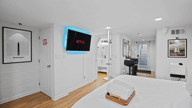 Una habitación de hotel moderna con una cama grande, un televisor montado mostrando 'NETFLIX', un cuadro enmarcado de Washington, y un baño visible a través de una puerta abierta.