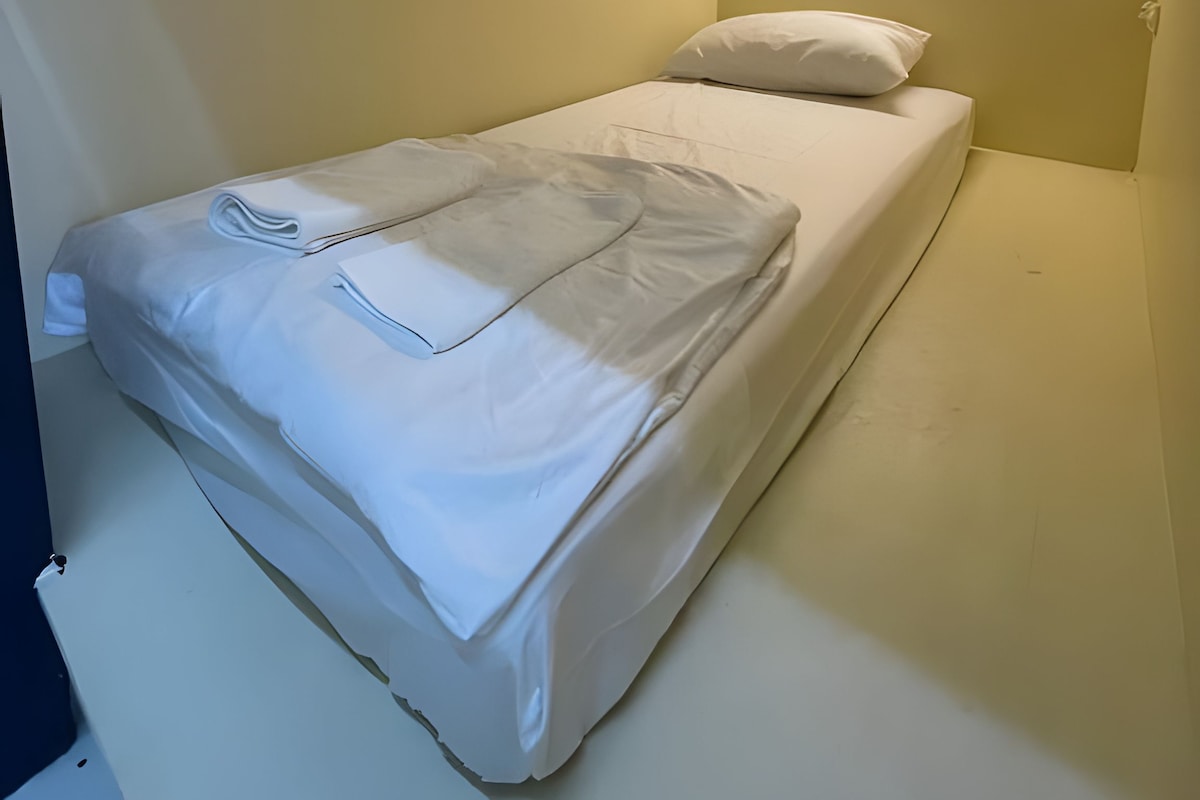 Een kleine kamer met een enkel bed, een effen witte dekbedovertrek en één kussen.