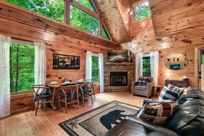 A cozy cabin interior with a dining area, fireplace, and a large window overlooking greenery.