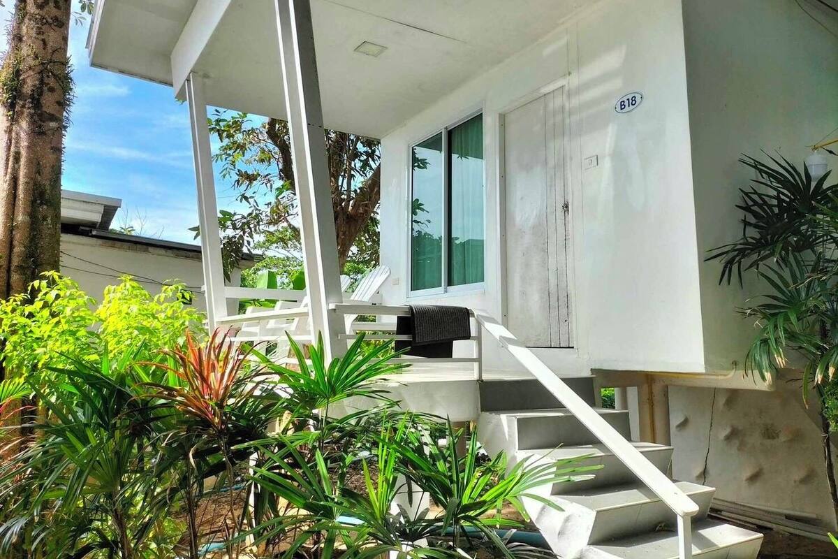 A modern house with a white exterior, a staircase leading to a covered porch, and lush greenery around.