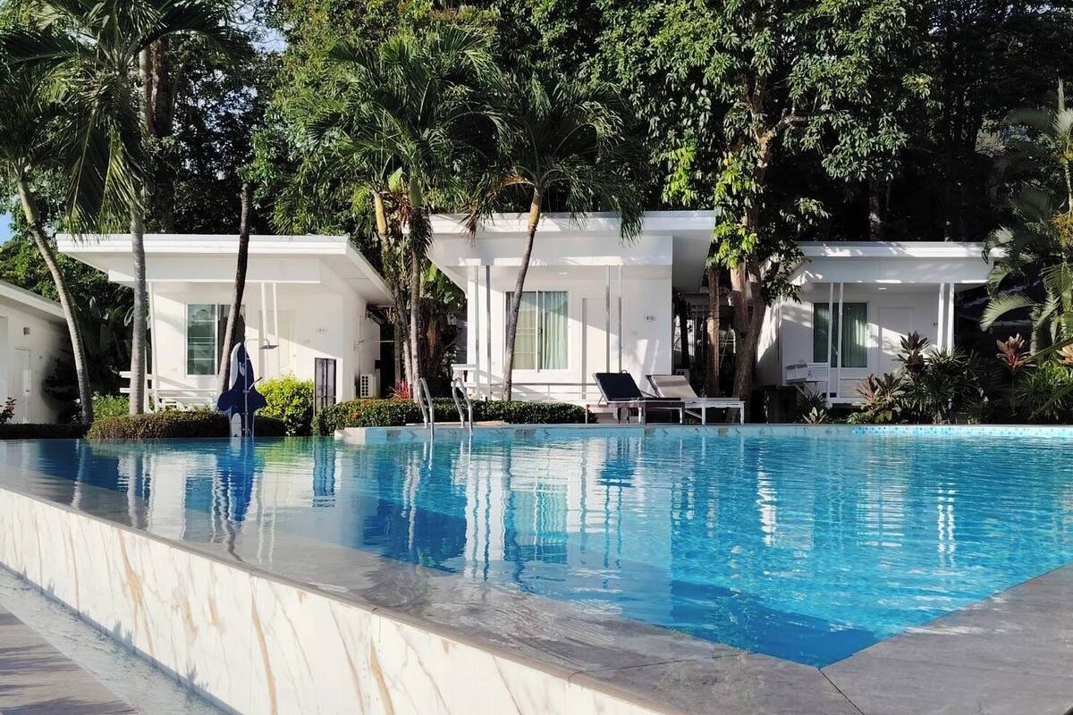 A swimming pool with lounge chairs, surrounded by palm trees and white buildings.