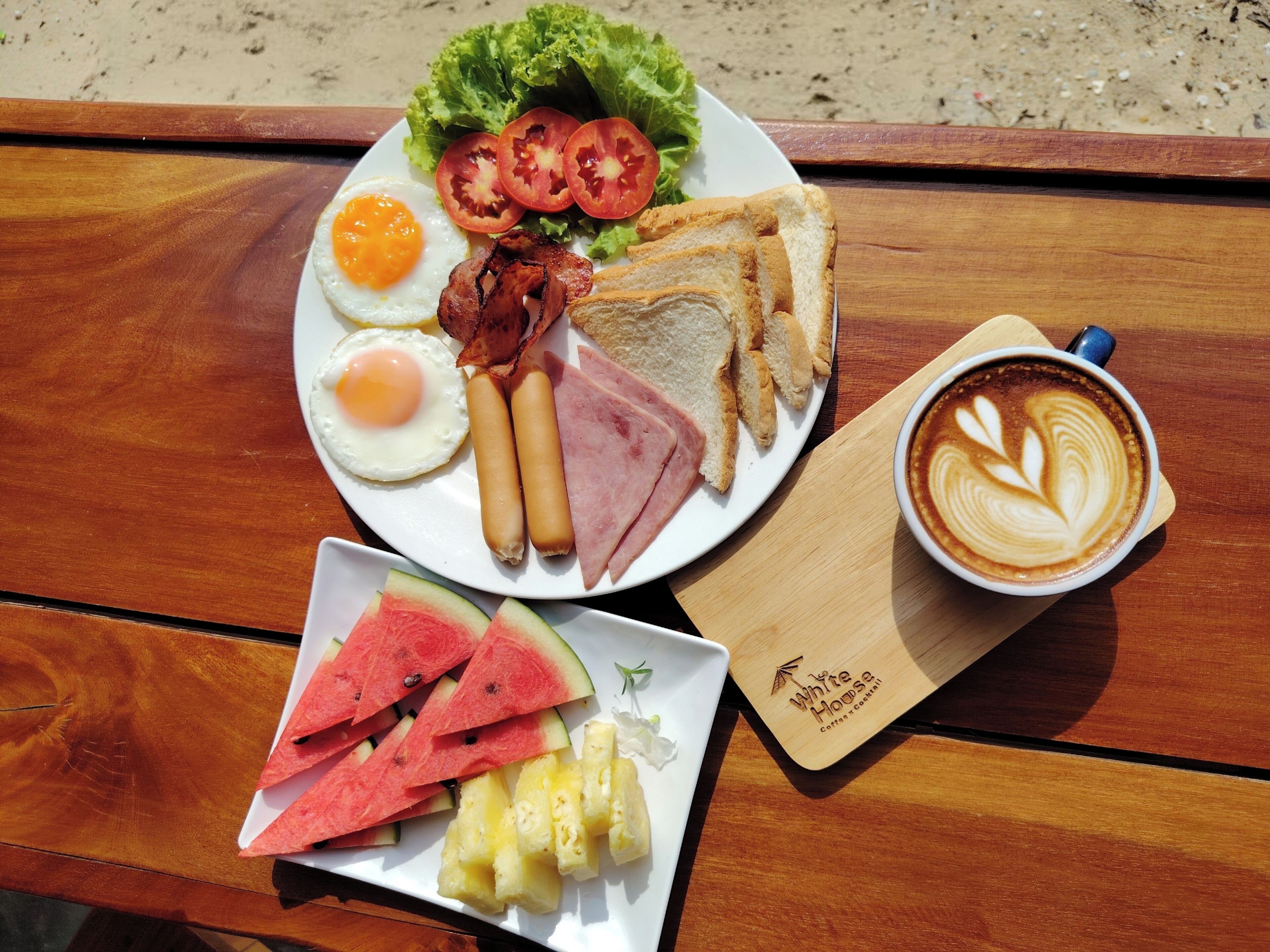 A plate of breakfast food including eggs, bacon, ham, toast, and tomatoes, along with a cup of coffee and a plate of watermelon and pineapple.