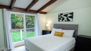 Een slaapkamer met een groot bed, een nachtkastje, een lamp en uitzicht op een groene tuin door een schuifdeur.