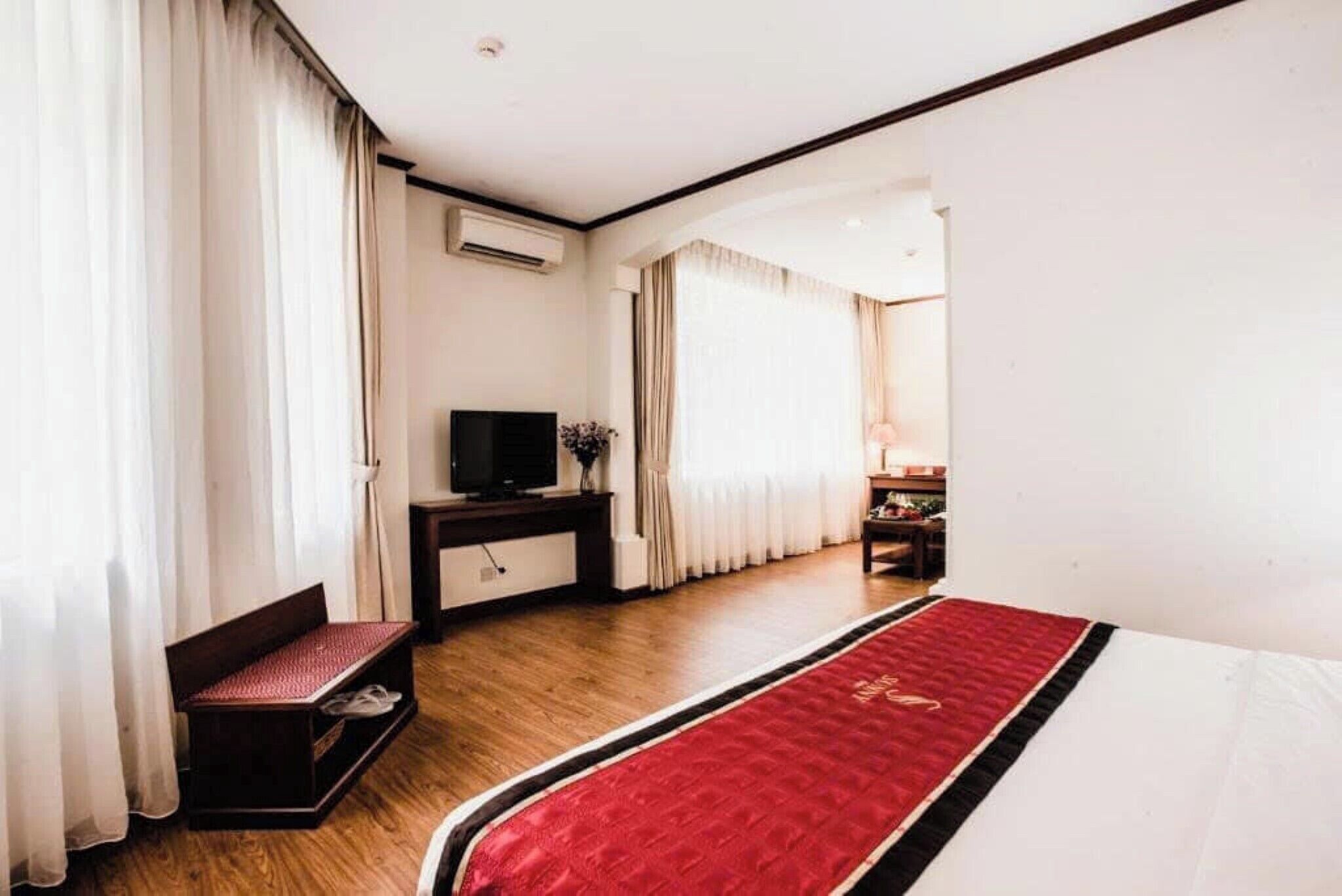 Une chambre d’hôtel avec un plancher en bois, un lit recouvert d’une couverture rouge, un téléviseur posé sur un meuble, un banc avec un coussin rouge et un bureau avec une lampe.