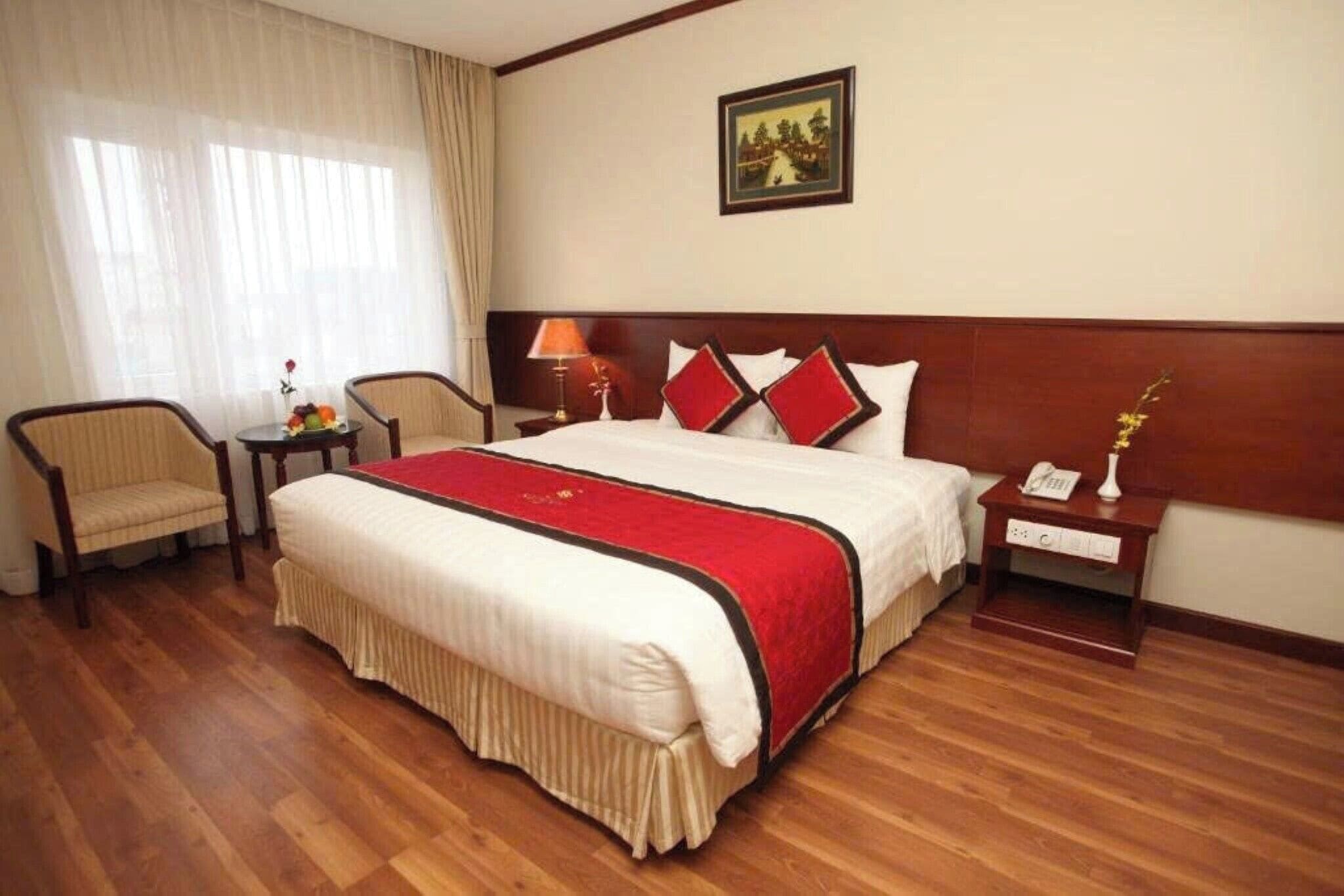 Une chambre d’hôtel avec un grand lit, deux tables de chevet, une petite table, une chaise et une fenêtre avec des rideaux.