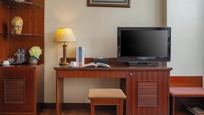 Un escritorio de madera con un televisor, una lámpara y libros. Hay un banco pequeño y una estantería con adornos.