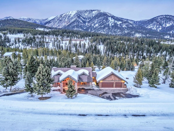 A large, multi-story lodge with a snow-covered roof, surrounded by a forest and mountains.