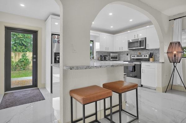 A modern kitchen with white cabinets, a granite countertop, and stainless steel appliances. There are two stools and a floor lamp.