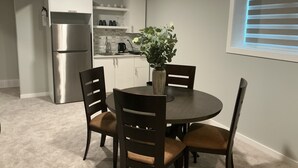 A compact kitchen with white cabinets, a stainless steel refrigerator, and a coffee maker. There is a dining table with four chairs and a vase with green plants.