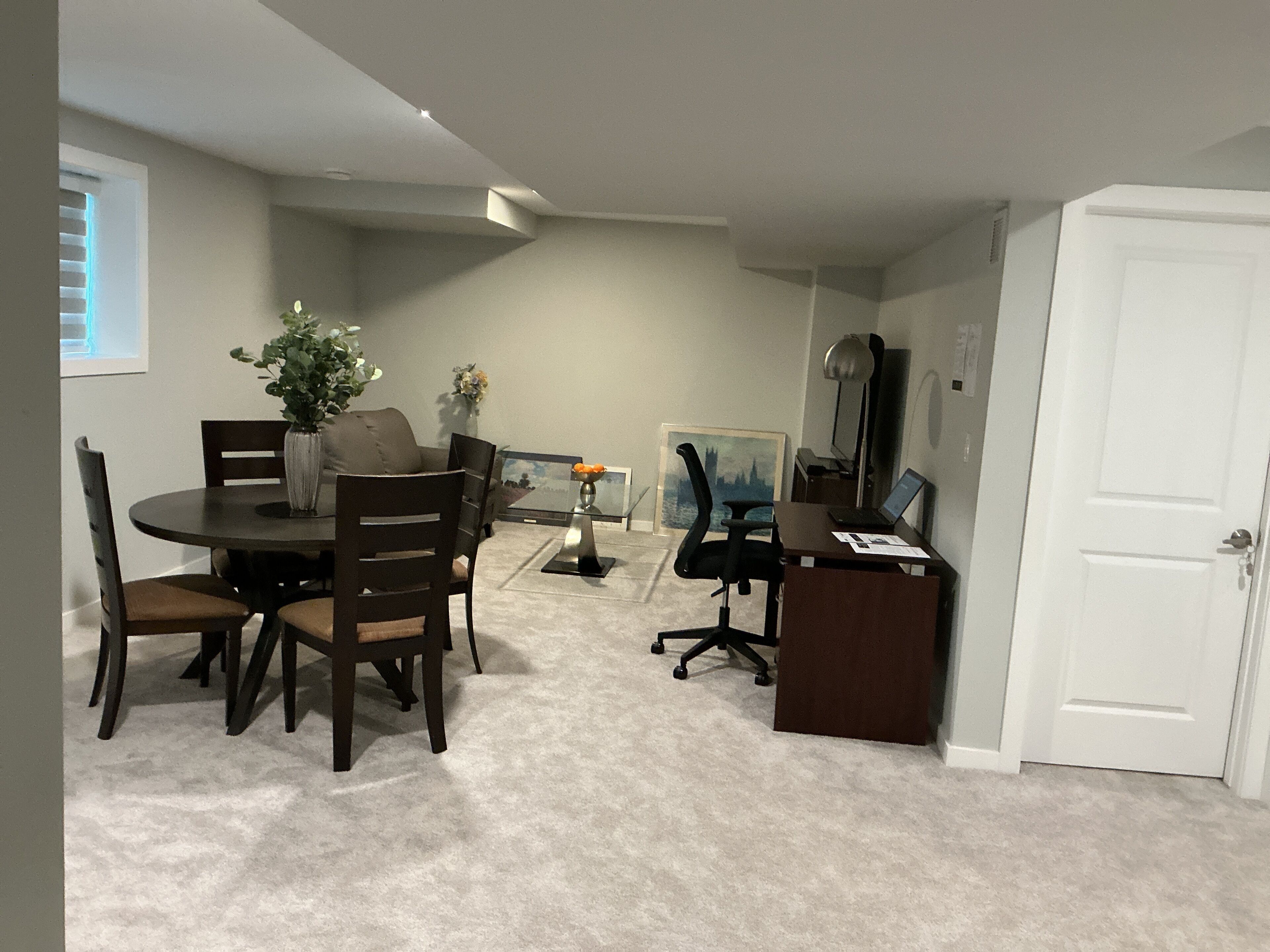 Un despacho pequeño con escritorio, computadora y silla de oficina. Se ve una mesa de comedor con sillas y un florero con flores. Un cuadro cuelga de la pared, y se observa una puerta.