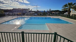 A rectangular swimming pool with a tiled deck, surrounded by a green fence, and a clear sky with scattered clouds. - Large 2-room luxury residence for 4 people with swimming pool and air conditioning. (LES ISSAMBRES)