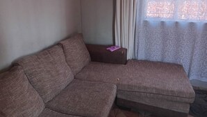 A living room with a brown sectional sofa, a small table with a purple object, and a window with patterned curtains. - Welcoming Bed & Breakfast with Air Conditioning in the Heart of Windhoek (Windhoek)