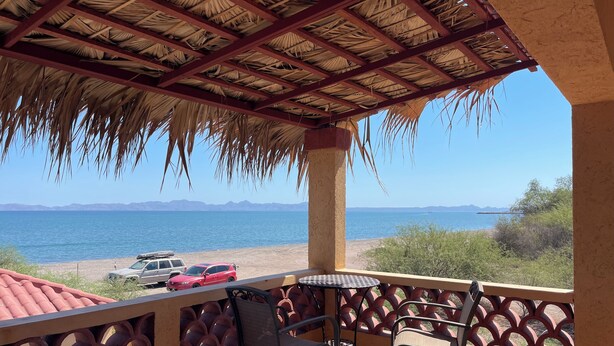 Un balcon doté d’un toit de chaume, donnant sur une plage où sont garées des voitures, et offrant une vue dégagée sur la mer et les montagnes lointaines.