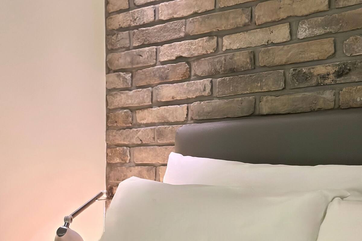 A bed with white bedding and a lamp beside it, against a brick wall.