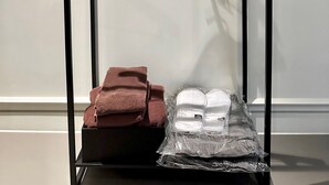 Un porte-manteaux en métal noir, muni de plusieurs cintres, est fixé contre un mur blanc. En dessous du porte-manteaux, on aperçoit une serviette marron et un sac en plastique contenant des articles blancs.