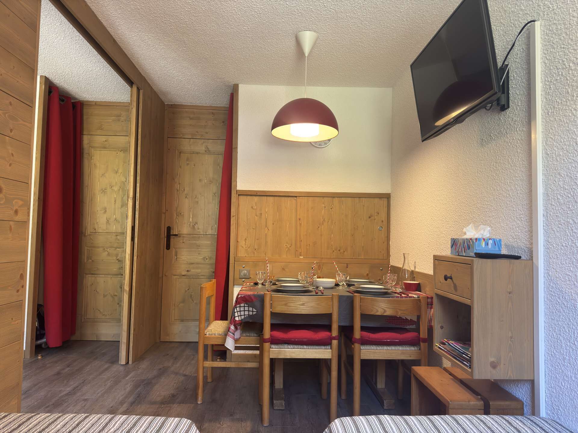 A wooden dining room with a table set for four, a television mounted on the wall, and a wooden cabinet with a shelf.