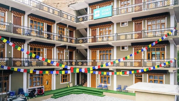 A multi-story hotel with a sign reading "Story Homes." It features balconies with railings, multiple windows, and a courtyard with chairs and a small table. - Story Homes- Deluxe Room (Kaza Khas)