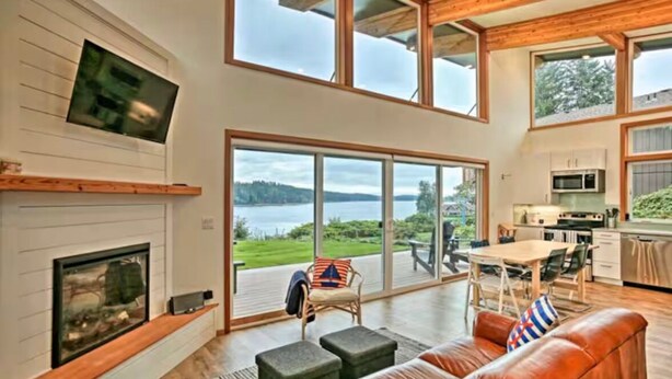 A spacious living room with a fireplace, a large window overlooking a lake, a dining area, and a kitchen.