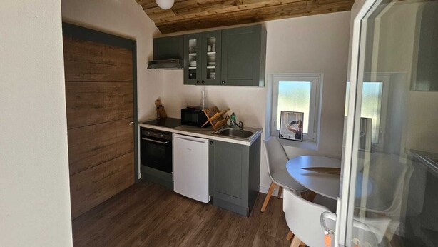 A compact kitchen with wooden ceiling beams, a dishwasher, and a small dining area with a table and chairs. - Furnished studio with all comforts (Beaumont-en-Véron)