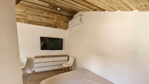 A bedroom with a wooden ceiling, a flat-screen TV mounted on the wall, a bed with a white duvet, a white chair, and a wooden dresser. - Furnished studio with all comforts (Beaumont-en-Véron)