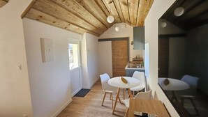 A modern kitchen with wooden ceiling beams, a dining area with white chairs, and a small table with a remote control. - Furnished studio with all comforts (Beaumont-en-Véron)