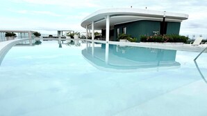 Area piscina moderna con acqua cristallina, poltrone prendisole e un edificio con tetto curvo.