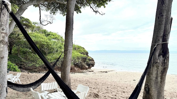Un'amaca tra gli alberi su una spiaggia sabbiosa, con poltrone da spiaggia bianche e vista sul mare.