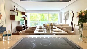 A modern kitchen with an induction cooktop, a dining area with a long table set for a meal, and a large window with a view of greenery. - Bolongo 3BR Your Exclusive Oceanfront Escape (Punta de Mita)