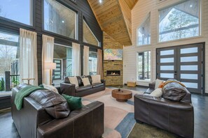 A spacious living room with leather sofas, a fireplace, large windows, and a garage door. - NEW! Creekfront, Pickleball, Hot Tub, Fire Pit, Forest, Hochatown, Beavers Bend (Broken Bow)