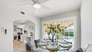 A modern dining area with a glass table set for six, surrounded by clear chairs. A ceiling fan is visible above, and a kitchen is seen in the background.