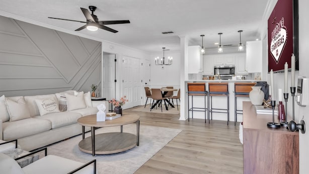 Una sala moderna con sofá, mesa de centro y ventilador de techo. Hay una zona de cocina con taburetes de bar y una mesa de comedor.