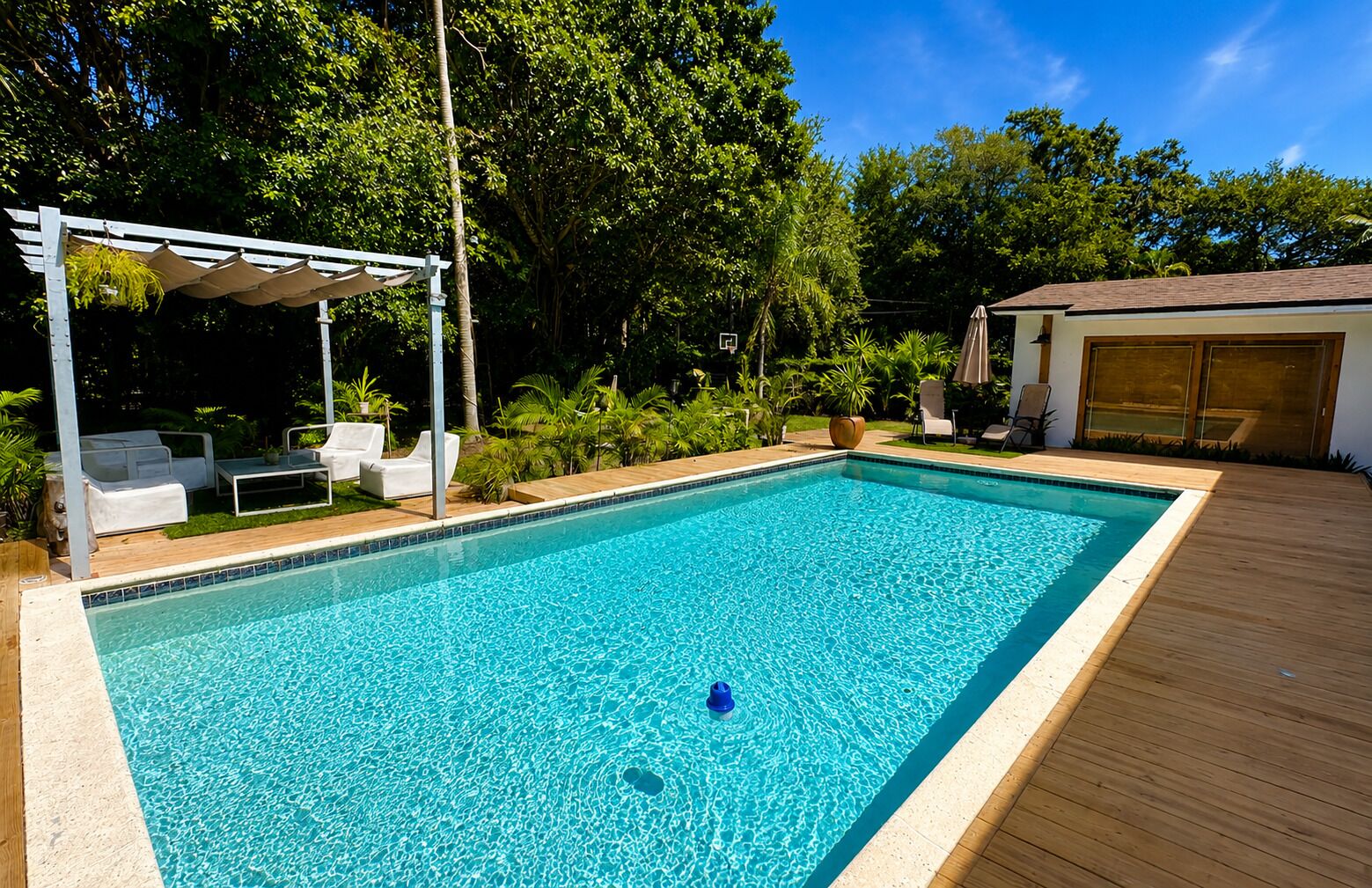 A rectangular swimming pool with a wooden deck surrounding it, a covered seating area with white lounge chairs, and a building with a garage in the background.