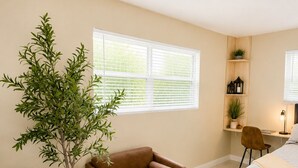 Quarto moderno com cama, poltrona marrom, mesa de centro com controles remotos, planta em vaso e escrivaninha com abajur.