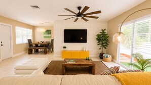 Uma sala de estar com sofá, mesa de centro, TV fixada em uma parede de tijolos brancos, ventilador de teto e um espaço de refeições com mesa e cadeiras.