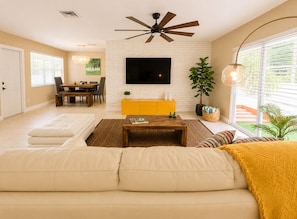 A living room with a sofa, a coffee table, a TV mounted on a white brick wall, a ceiling fan, and a dining area with a table and chairs.