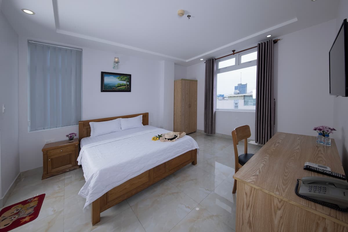 Une chambre d’hôtel avec un lit en bois, un bureau avec un téléphone, une chaise, une fenêtre avec des rideaux, une armoire et un tableau au mur.
