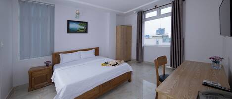 A hotel room with a wooden bed, a desk with a telephone, a chair, a window with curtains, a wardrobe, and a painting on the wall.