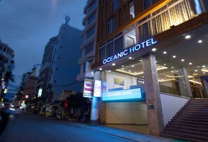 La façade de l’hôtel Oceanic, avec une enseigne bien visible, un escalier menant à l’entrée et une vue sur la rue, où l’on aperçoit d’autres bâtiments et des panneaux.