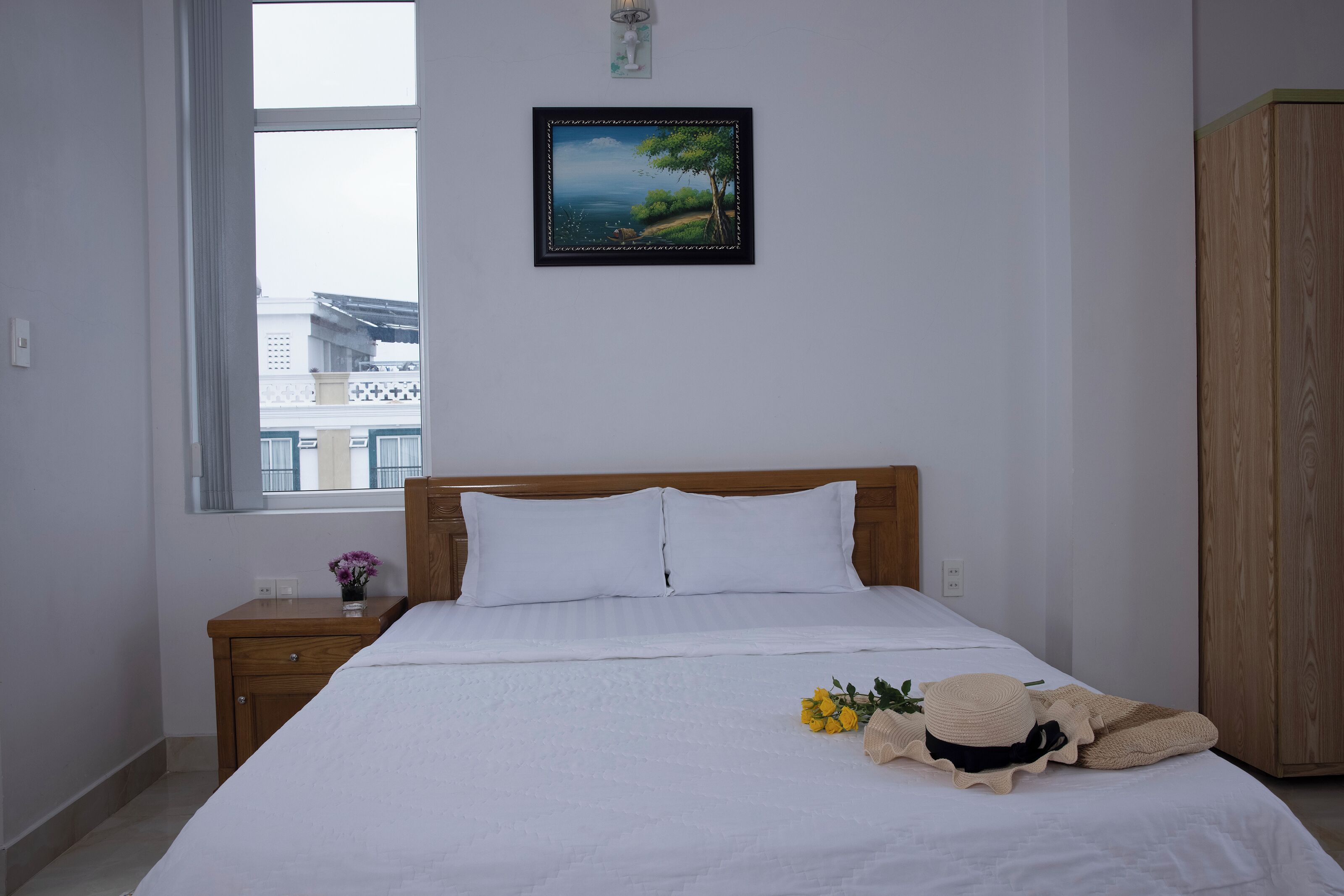 A neatly made bed with white bedding, a wooden headboard, a nightstand with a small vase of flowers, and a framed picture of a landscape on the wall.