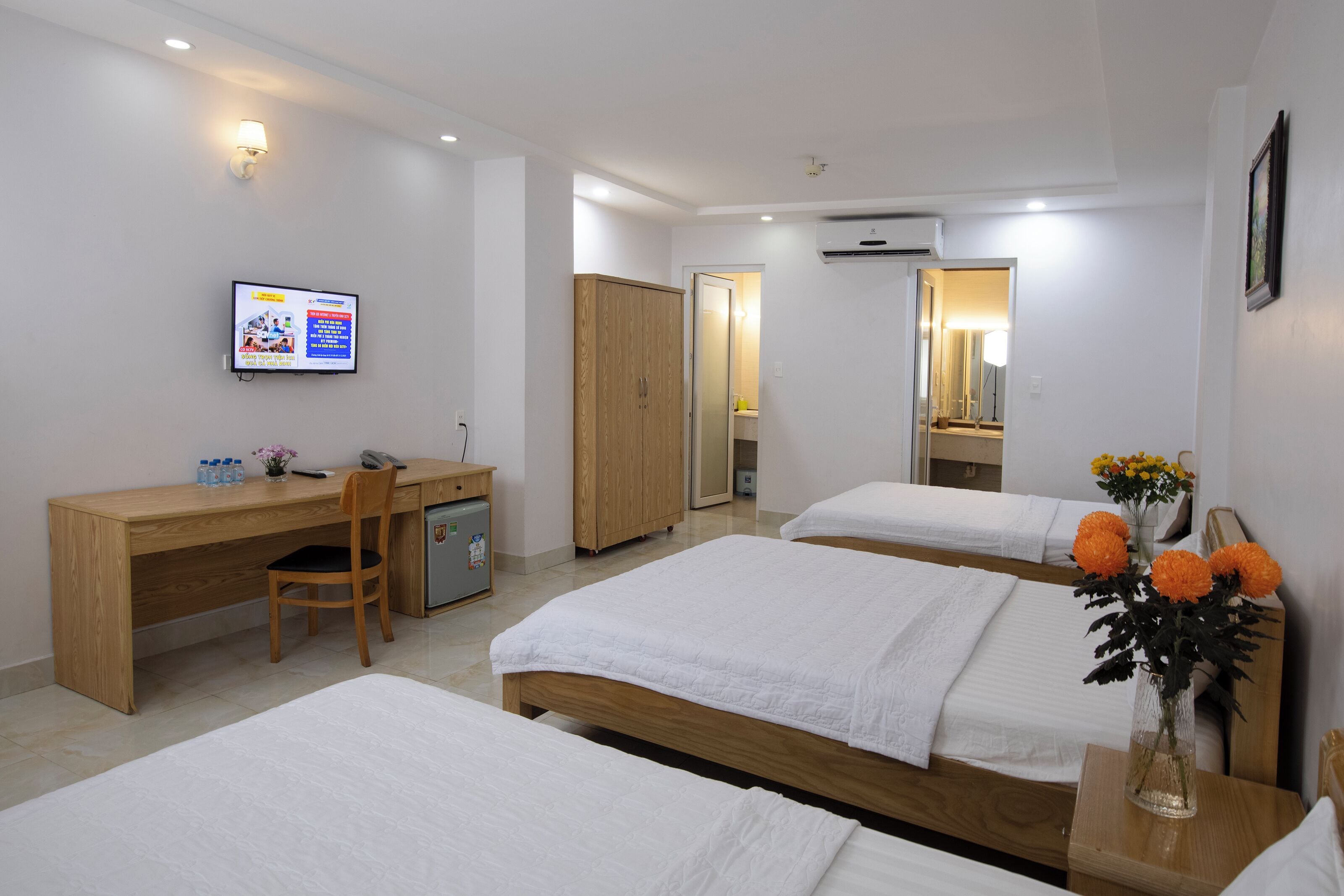 Une chambre d’hôtel avec un bureau en bois, deux lits simples, un petit réfrigérateur, une télévision et un vase avec des fleurs.
