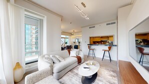 Un salon moderne avec un canapé, une table basse et des tabourets de bar. On aperçoit un coin repas avec des chaises et, au fond, une cuisine.