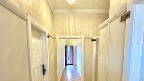 Un pasillo con paredes blancas, suelo de madera en dibujo de espiga y una araña. Puertas a la izquierda y a la derecha dan acceso a otras habitaciones.