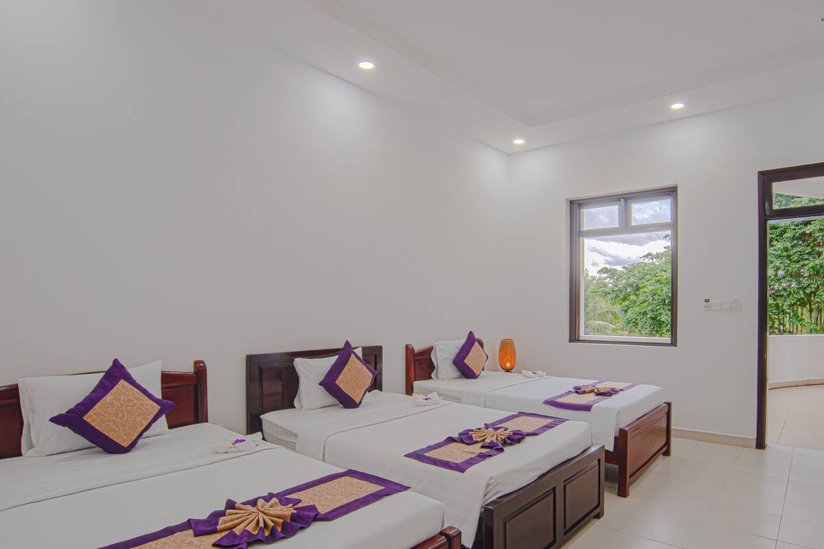 A hotel room with three single beds, each with a white and purple bedspread, and a large window providing a view of greenery outside.
