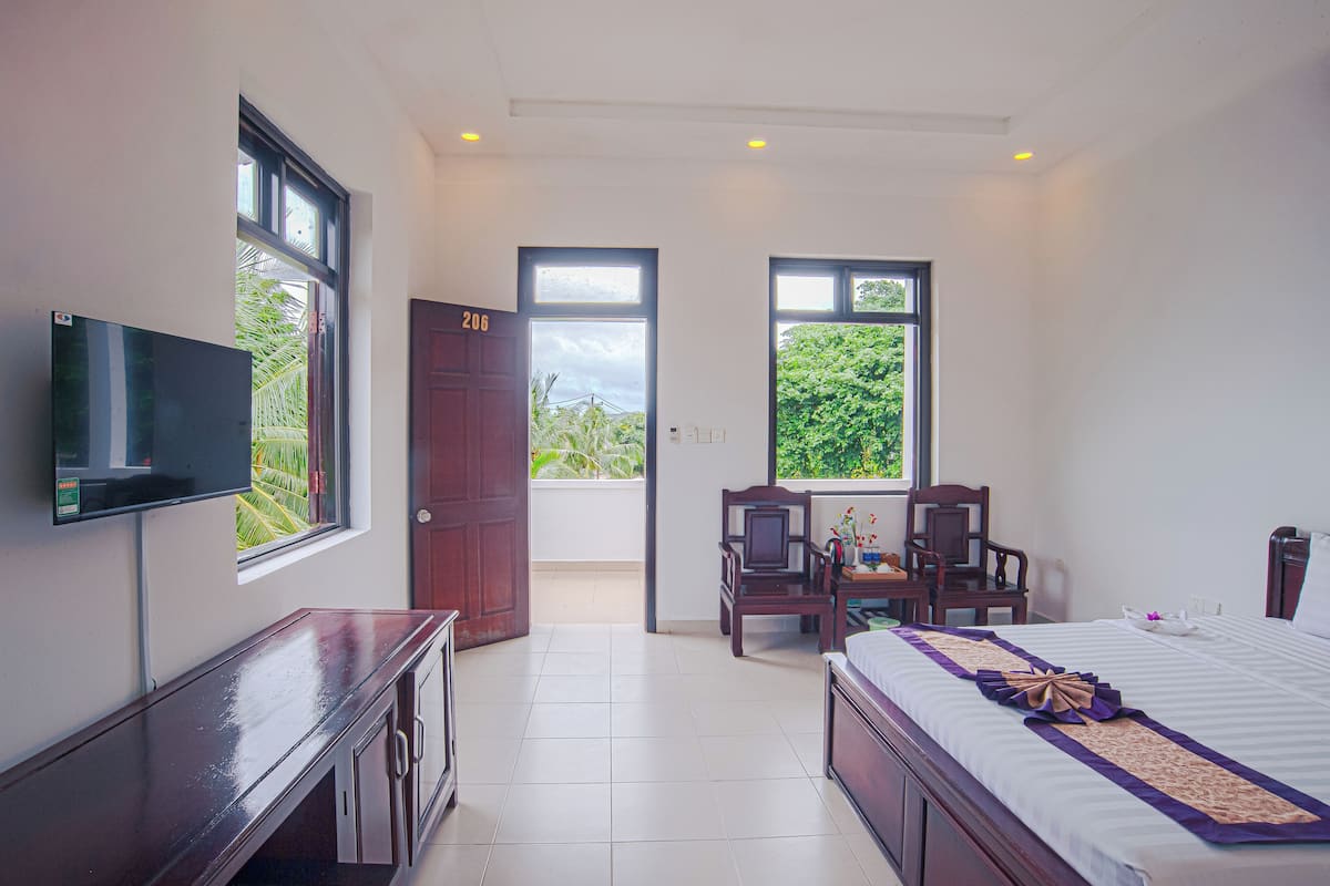 A hotel room with a bed, a desk, a TV, and two windows offering views of greenery.